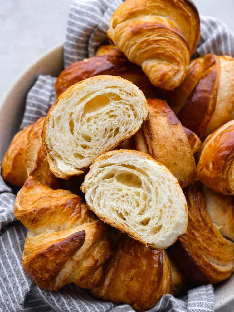 Close up view of croissants stacked  on a gray striped kitchen towel and  in a large cream colored bowl. One is cut and separated down the middle to show the baked inside layers.