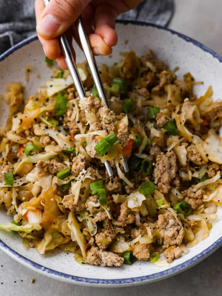 A pair of chopsticks getting some eggs roll in a bowl out of the white bowl that the food is in. 