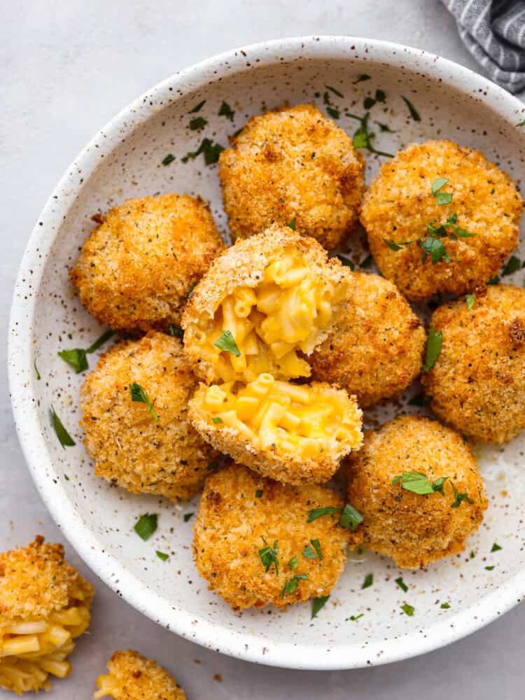 A speckles, white bowl filled with Mac and cheese balls. One of them is broken open so that you can see the inside. 