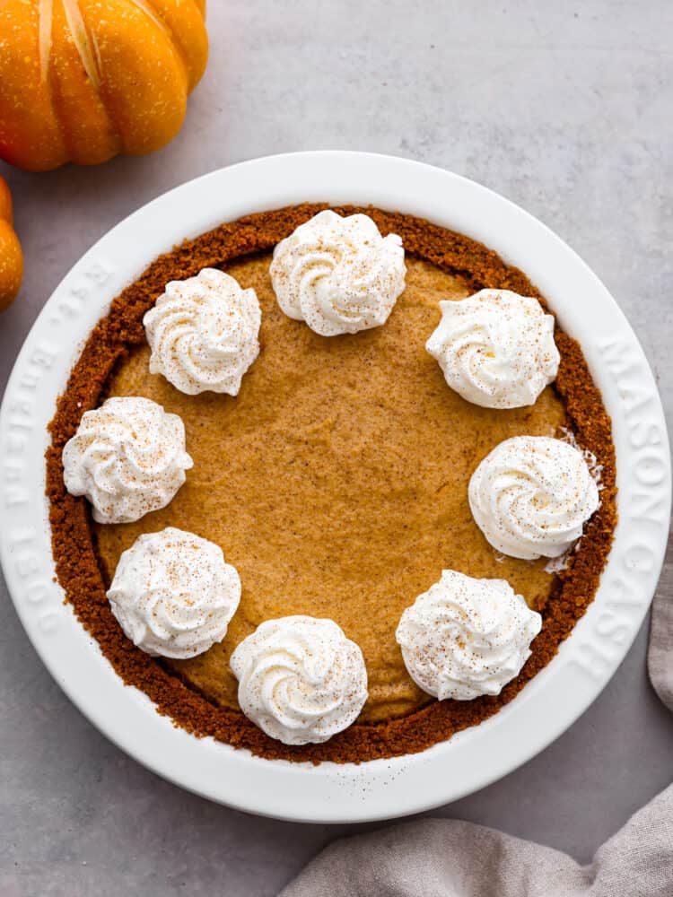 An overhead shot of Pumpkin Chiffon pie in a white pie dish with whipped cream and dusted with cinnamon next to a tea towel and small pumpkins.