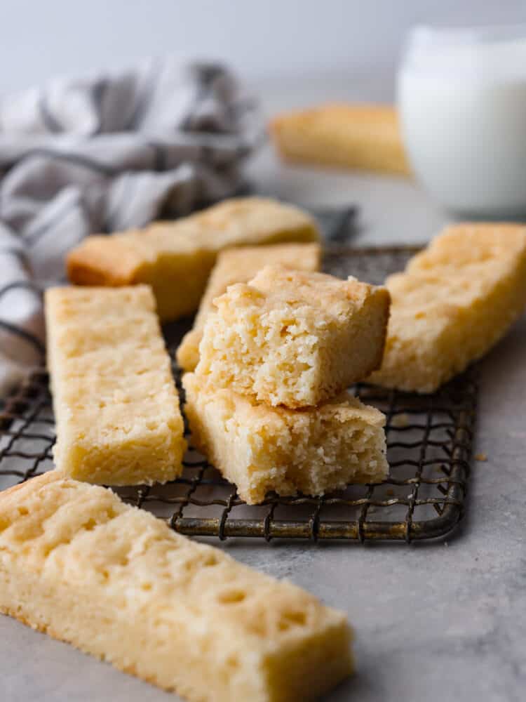 Close up view of Scottish shortbread stacked on a cooling rack.  One cookie is broken into two pieces. A kitchen towel and glass of milk are in the distance.