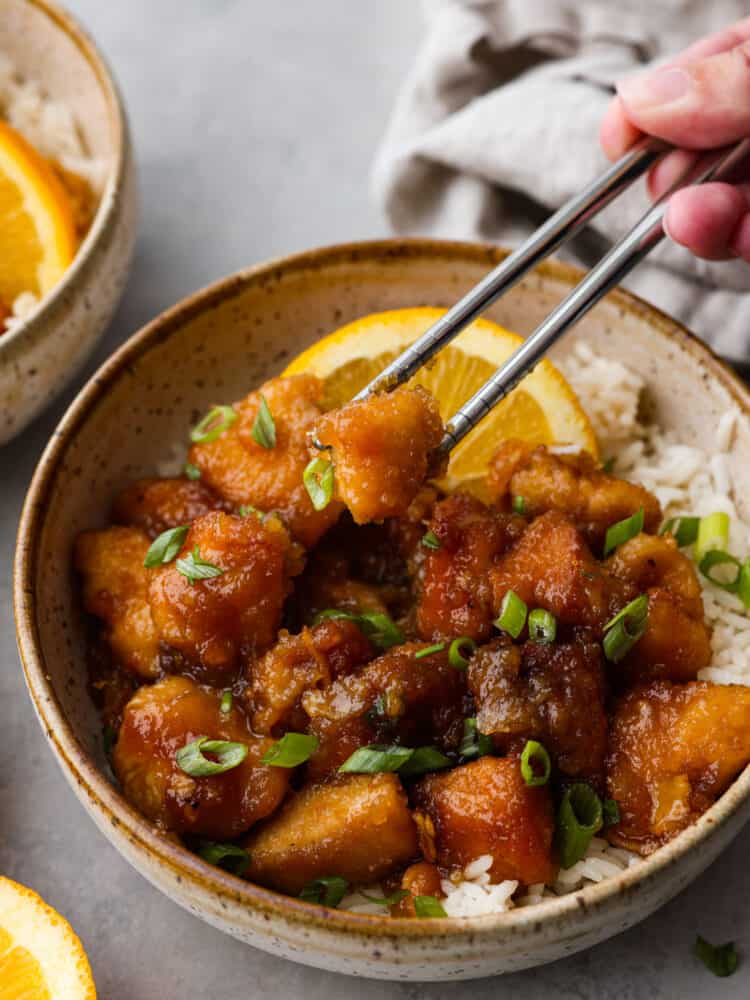 Orange chicken in a bowl with some silver chopsticks taking out a piece. 