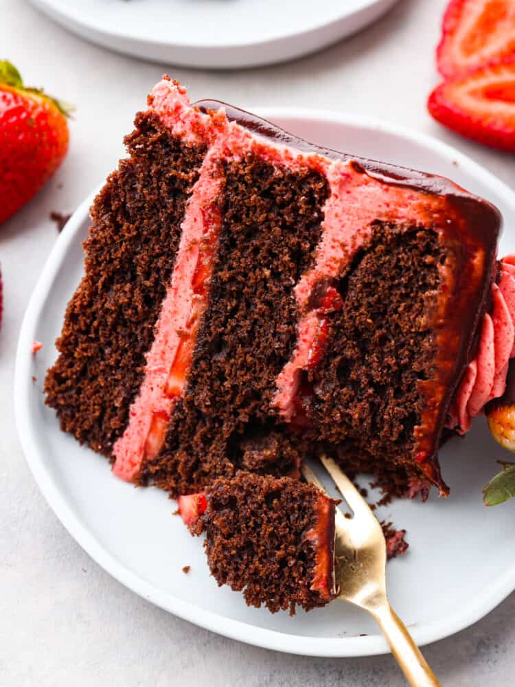 A close up photo of a slice of strawberry chocolate cake on a gray plate.  A fork is lifting up a bite of cake. Sliced strawberries are garnished on the side of the plate.