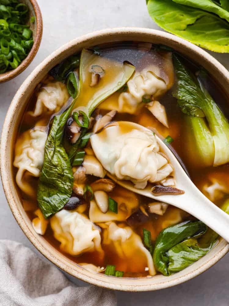 Top view of wonton soup in a tan bowl with a white spoon lifting up a wonton dumpling.  A tan towel and bowl of green onions are next to the bowl.