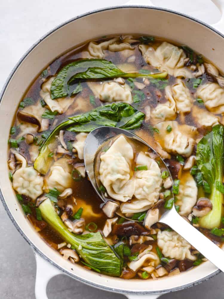 Large white pot full of wonton soup.  A large silver serving spoon is lifting the soup out of the pot.