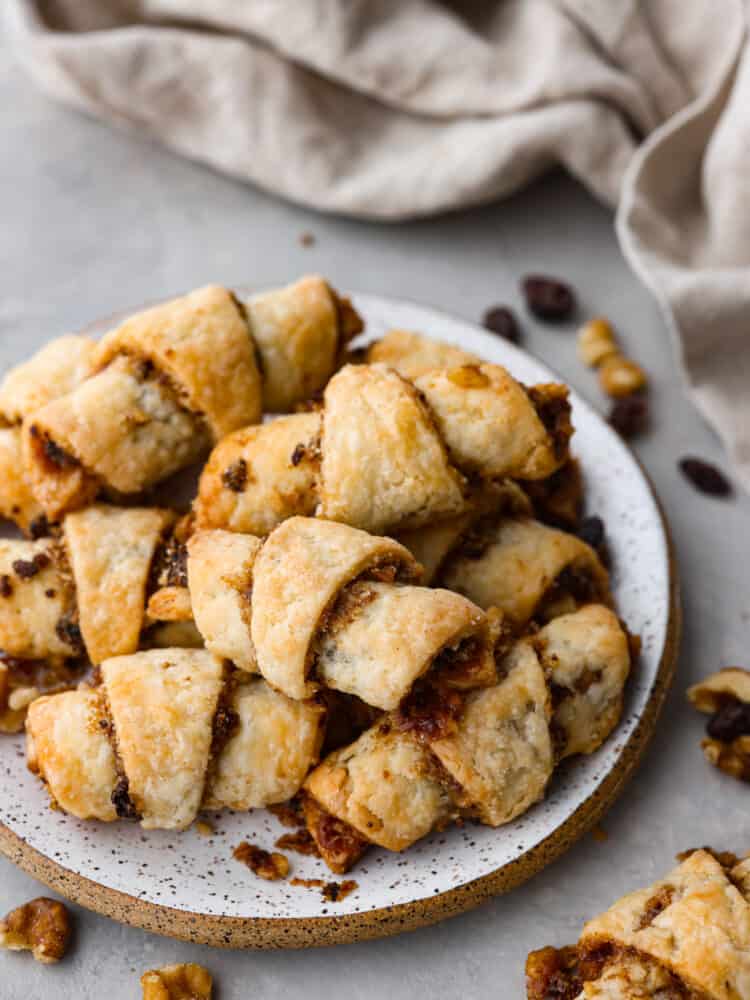 A closeup photo of a small plate of rugelach cookies stacked on top of each other. Raisins, walnuts, and a kitchen towel are scattered around the plate.