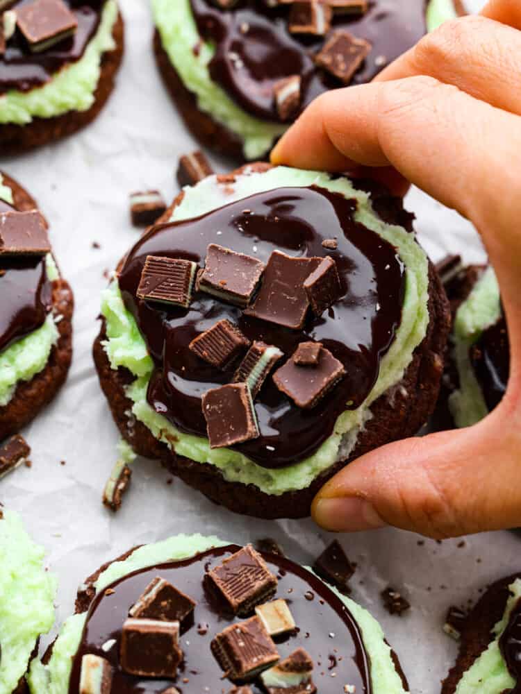 A hand holding a chocolate cookie with other cookies in the background.