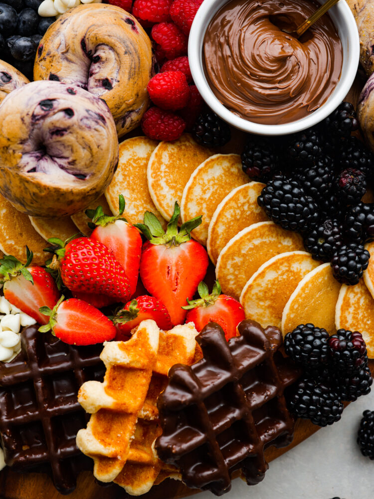 Close up view of pancakes, waffles, and bagels on the breakfast charcuterie board. Colorful fruit and Nutella are scattered around the pancakes.