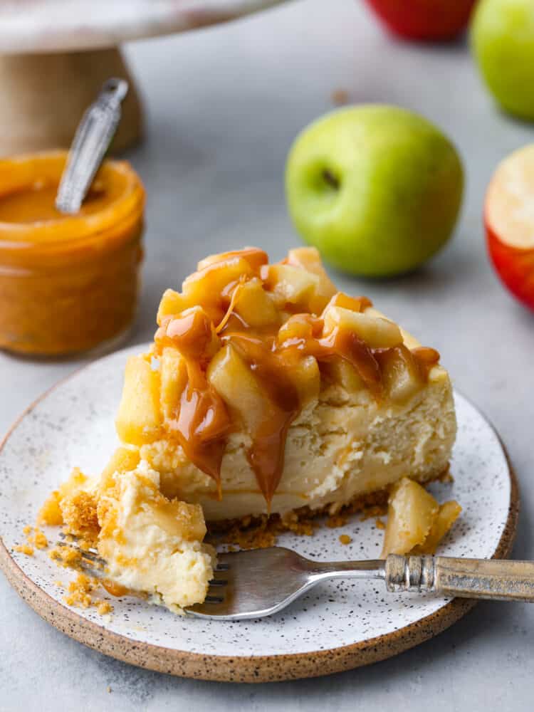 A slice of Caramel Apple Cheesecake with a bite taken out by a fork sitting on a stone ware plate with caramel and apples in the background.