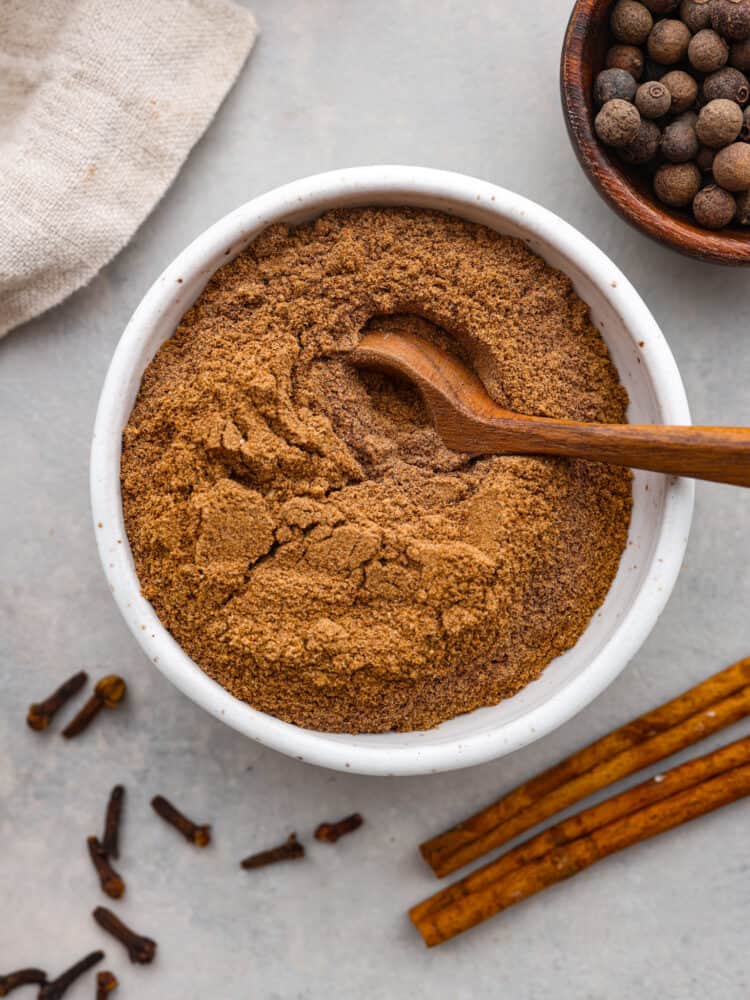 Chai spice in a white bowl with a wooden spoon sitting in it with spices around it.