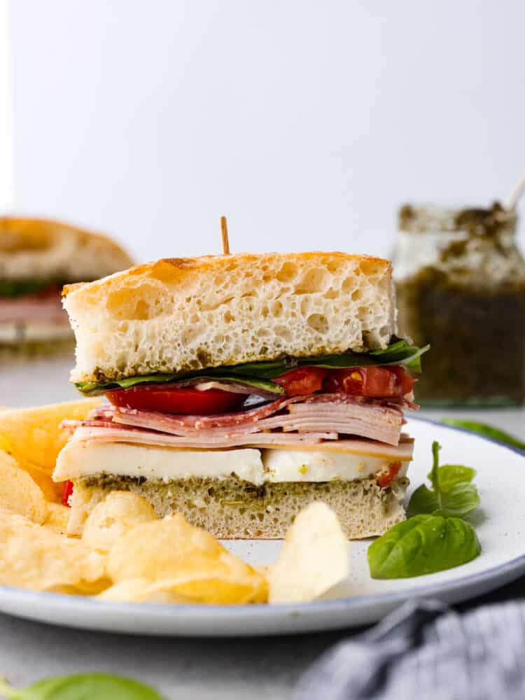 Ciabatta sandwich on a white plate with potato chips and basil next to it.