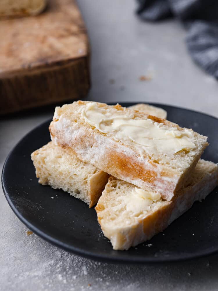 Close up view of sliced and buttered bread on a black plate. A wood cutting board nearby with scattered crumbs around the plate.