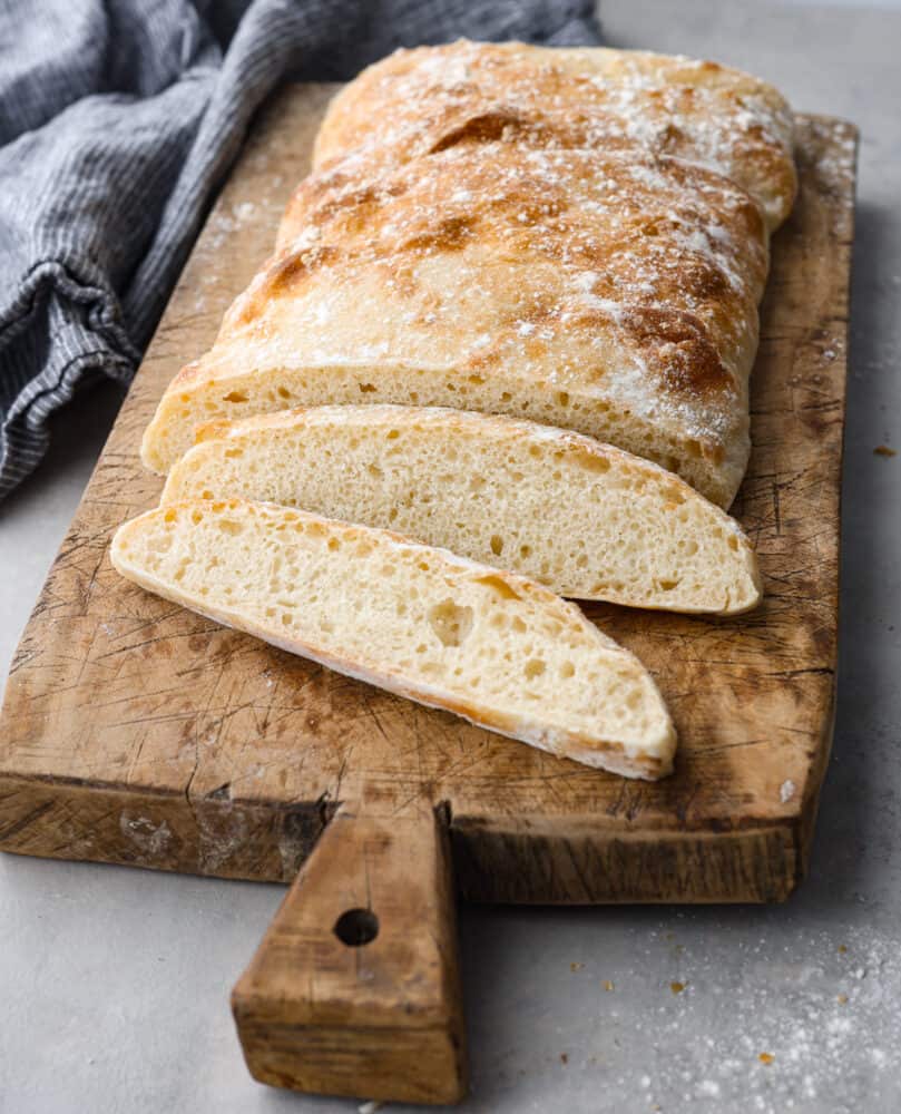 Side view of baked and slice ciabatta bread on a wood cutting board. A blue kitchen towel is next to the wood board.