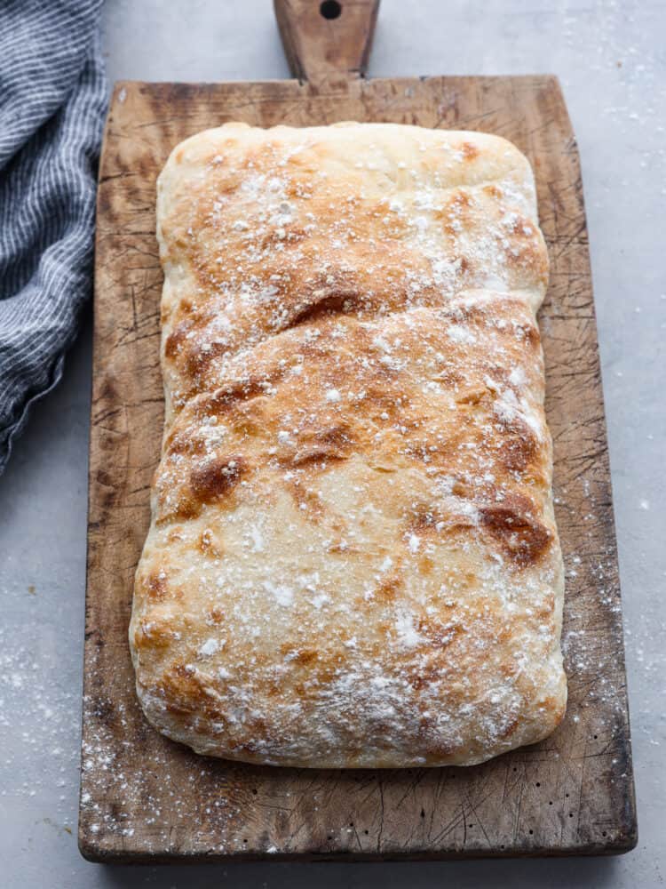 Top view of a loaf of ciabatta bread on a wood cutting board. A blue kitchen towel is next to the wood board. Flour is sprinkled on top of the loaf.