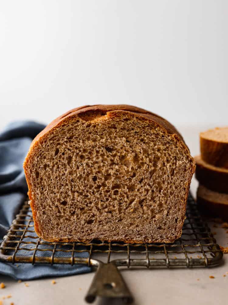 Whole wheat bread loaf sliced open facing the viewer sitting on a wire rack with stacked bread in the background.