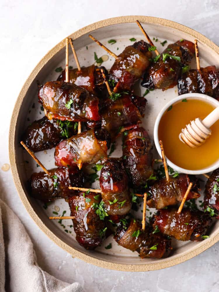 Top view of bacon wrapped dates on a tan colored plate.  A small bowl of honey is on the plate with a tan colored kitchen towel styled next to the plate.