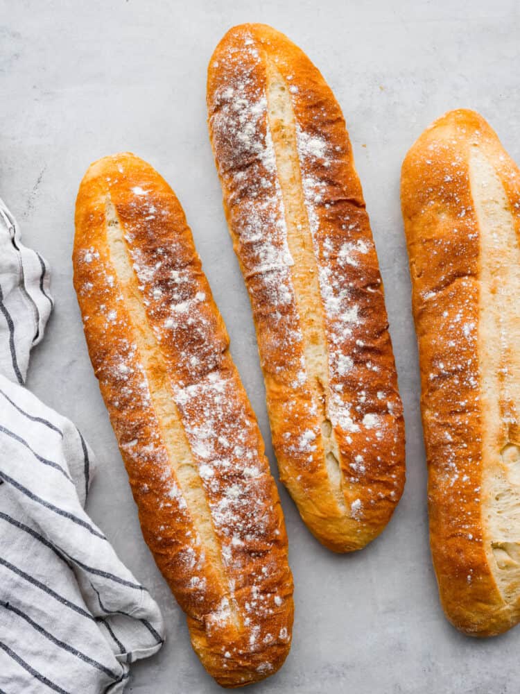 Three loaves of cuban bread sitting on a counter top with a tea towel next to them.