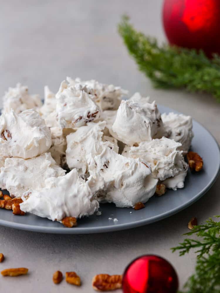 A pile of divinity on a gray plate. Pecans, greenery, and ornaments are styled around the plate.