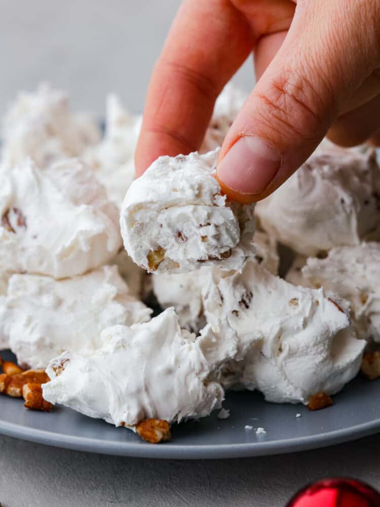 Close up view of a hand holding divinity with a bite out of it. Pecans are scattered around the plate of divinity.