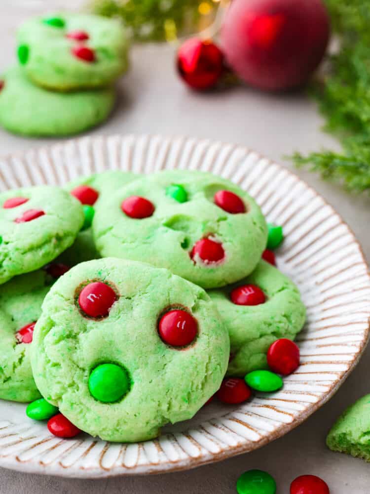 Close up view of Grinch cookies stacked on a cream colored plate. Christmas ornaments, greenery and extra cookies are styled next to the plate.