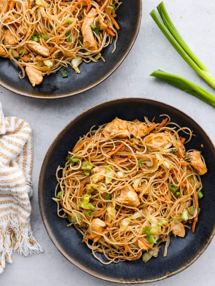 Top view of pancit noodles into two black bowls. Green onion and a tan kitchen towel are next to the bowls.