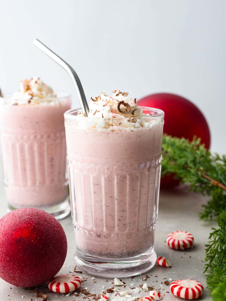 2 glasses of peppermint milkshakes surrounded by red Christmas ornaments and  pine tree branches.