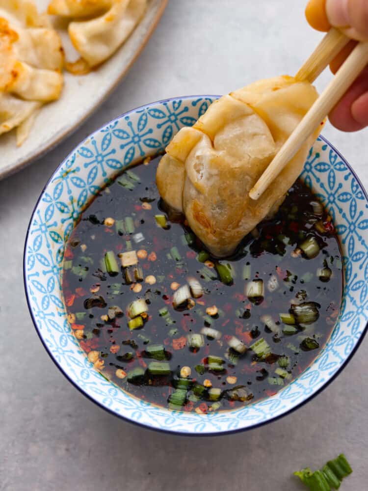 Close up view of potsticker sauce in a blue bowl. A potsticker is being dipped into the sauce with chopsticks. 