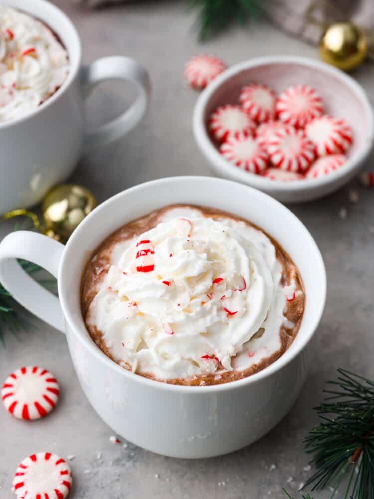 Peppermint hot chocolate in a white mug, topped with whipped cream and crushed peppermints. There is a dish filled with peppermints in the background.