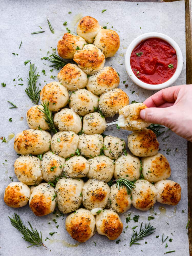 Christmas tree pull apart bread sitting on parchment with some sauce next to it and a hand pulling away some of the bread.