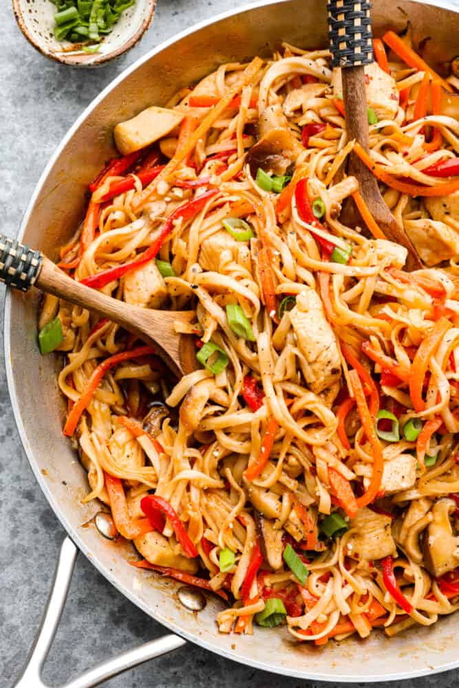 A close up on chicken lo mein in the skillet with a wooden spoon in it.