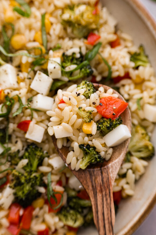 Orzo pasta salad on a wooden spoon with a bowl in the background.