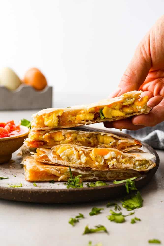 Close up view of a breakfast quesadilla cut and stacked on a brown plate. Chopped cilantro and salsa are on the plate. Fresh eggs are styled next to the plate.