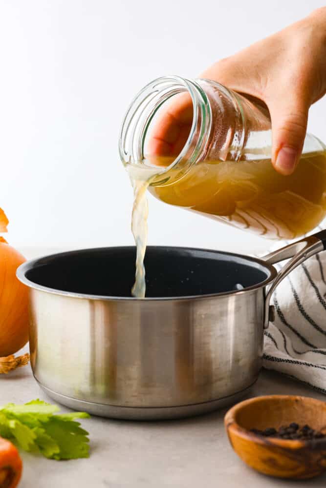 Side view of chicken broth being poured into a silver pot. Vegetables and peppercorns are next to the pot.