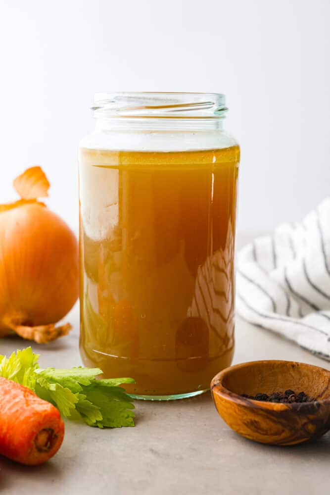 Close up view of chicken broth in a large. mason jar with onion, celery, and carrots scattered next to the jar.