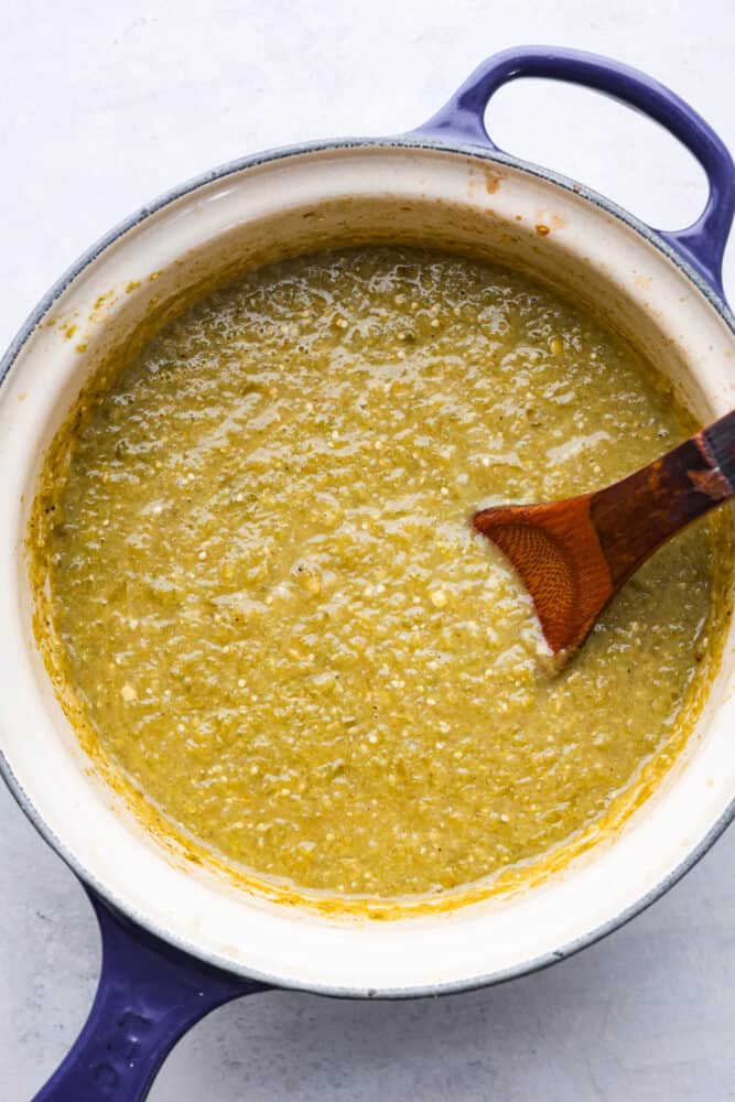 A blue pot with a wooden spoon filled with cooked green enchilada sauce 