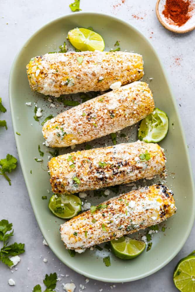 A sage green platter with 4 ears of corn slathered with the mayonnaise mixture and sprinkles with spices and cheese. Lemon wedges garnish the plate. 