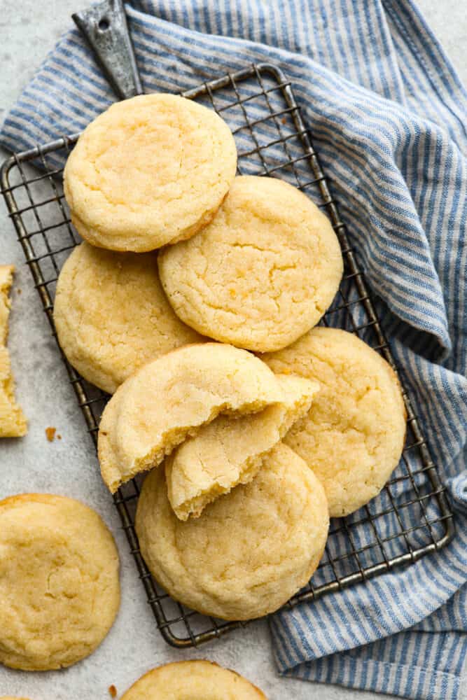 Close up side view of soft sugar cookies stacked on a cooling rack. The cookie on top of the stack is broken in half with the other half on the counter. 