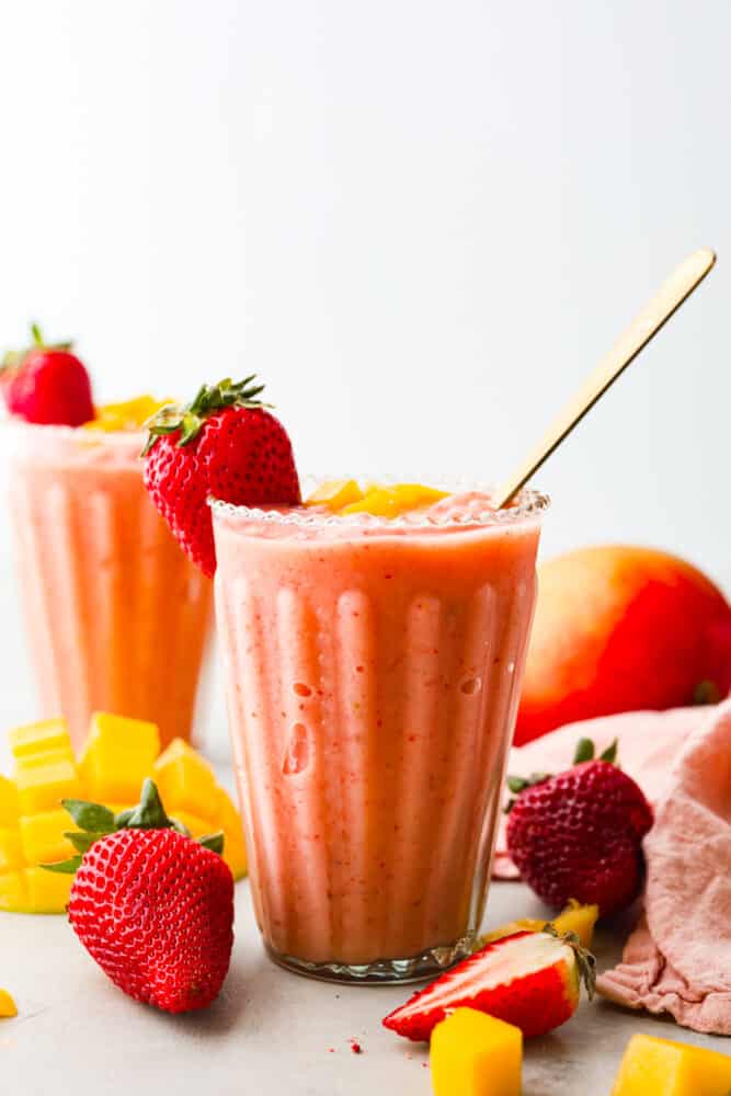 A strawberry mango smoothie in a clear glass with a gold spoon. Strawberries and mangoes are around the cup. 