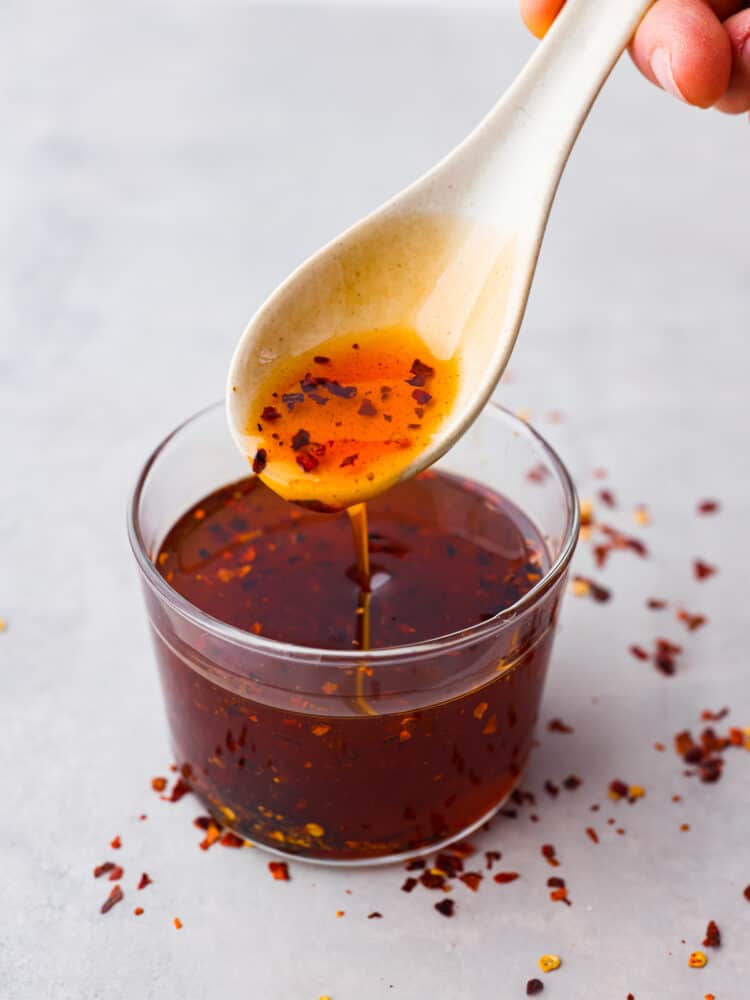 Top view of chili oil being spooned out of a clear glass jar. Red pepper flakes are scattered on the table.