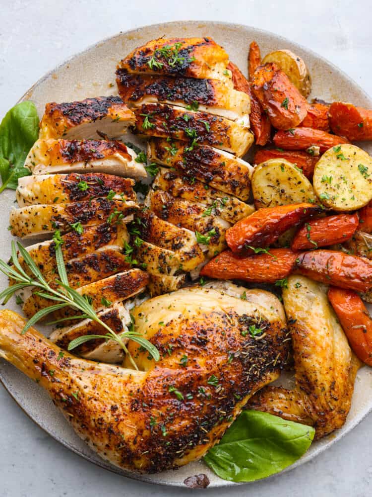 Close up view of spatchcock chicken carved and served on a platter with roasted carrots, potatoes, and fresh herbs.