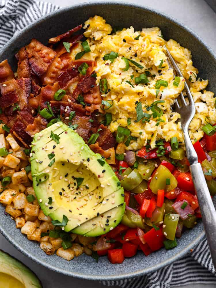 Close view of a breakfast bowl with a fork in the bowl. Eggs, cheese, bacon, potatoes, and vegetables are in the bowl with chopped cilantro on top.