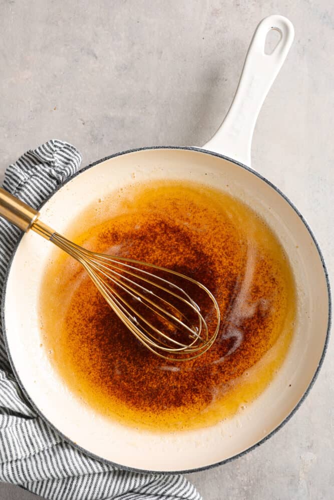 Top view of browned butter in a white skillet. A gold whisk is inside the skillet with a blue and white striped towel next to the skillet.