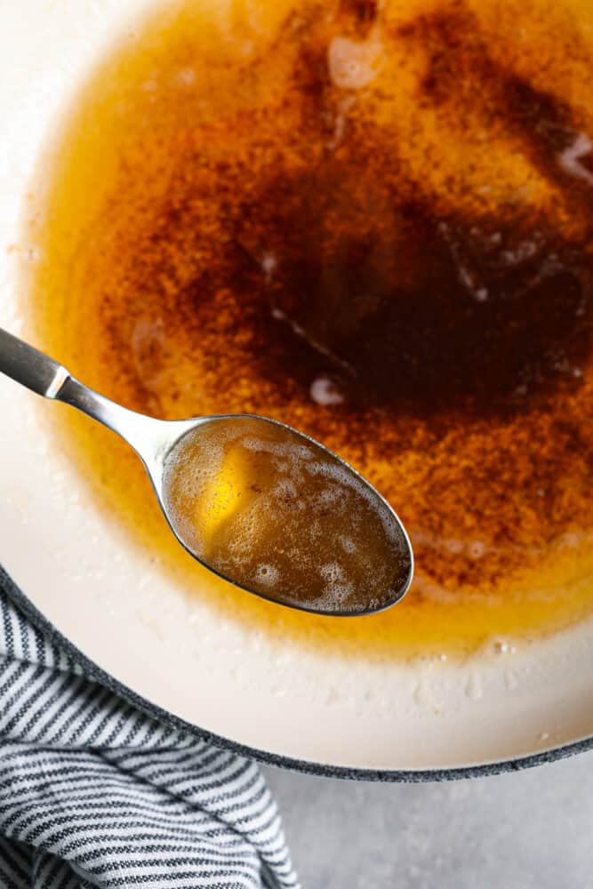 Close up view of a spoon lifting up brown butter out of a white skillet. A blue and white striped towel is next to the skillet. 