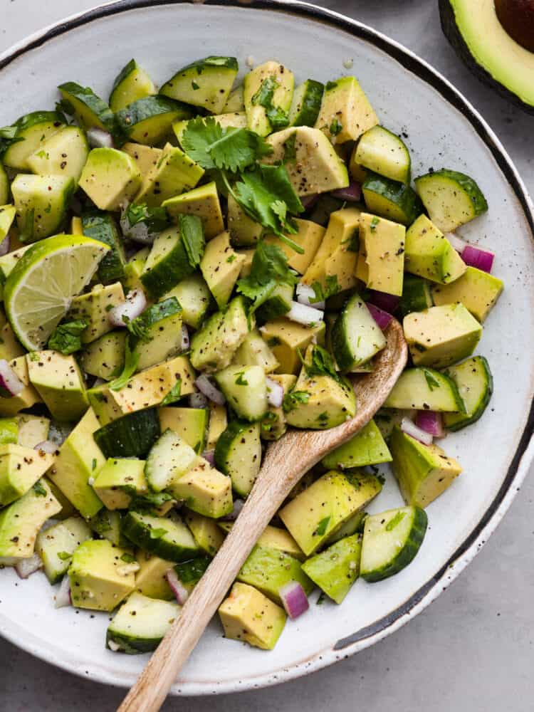 Cucumber avocado salad in a white bowl with a wooden spoon.