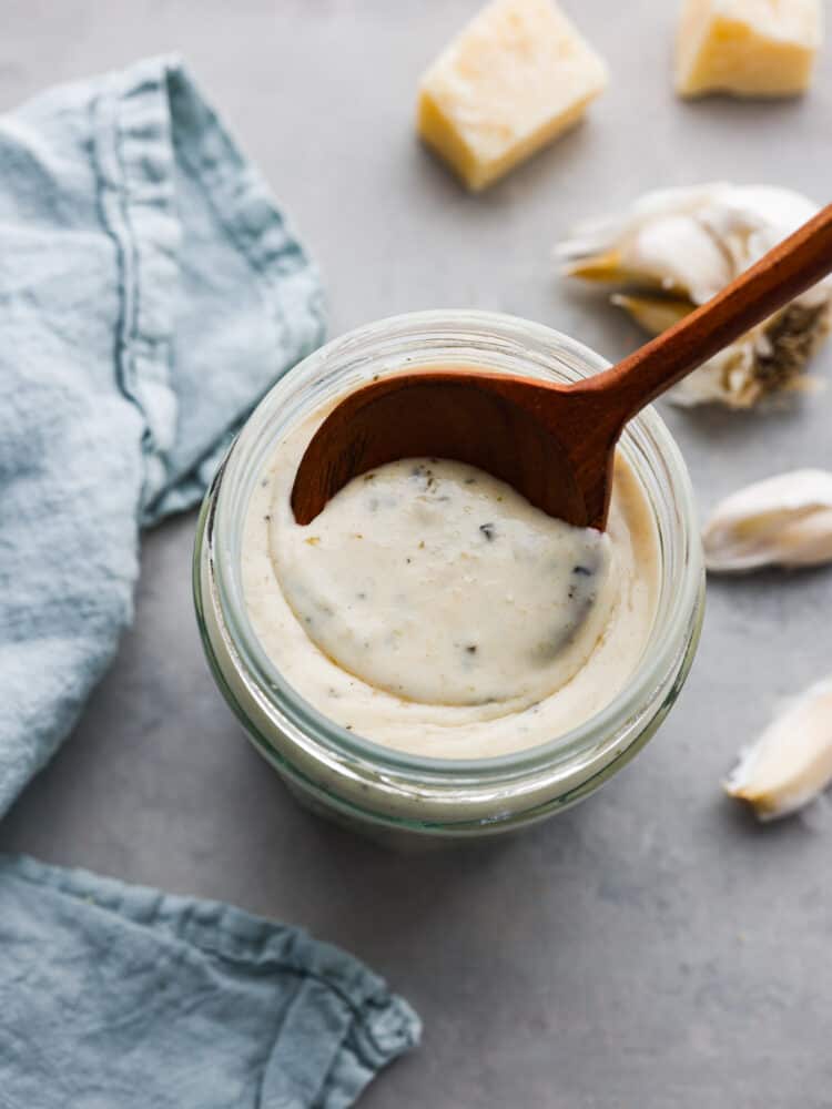 Close up view garlic parmesan sauce in a glass jar with a wood spoon in the jar. Fresh garlic and parmesan cheese next to the jar.