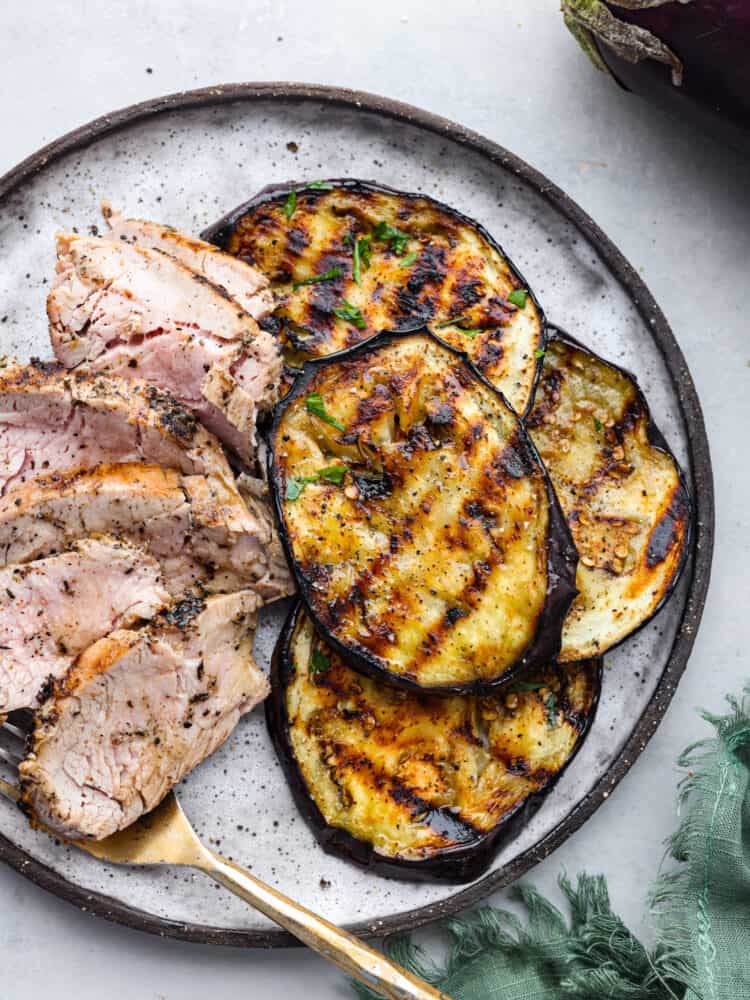 Top view of grilled eggplant on a white plate along with sliced pork and gold fork.