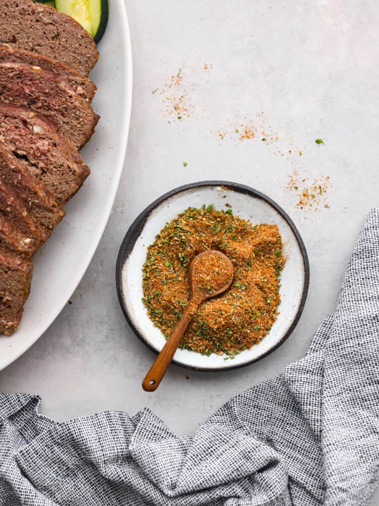 Top view of gyro seasoning in a white bowl with a wood spoon. A platter of gyro meat and a kitchen towel are next to the seasoning.