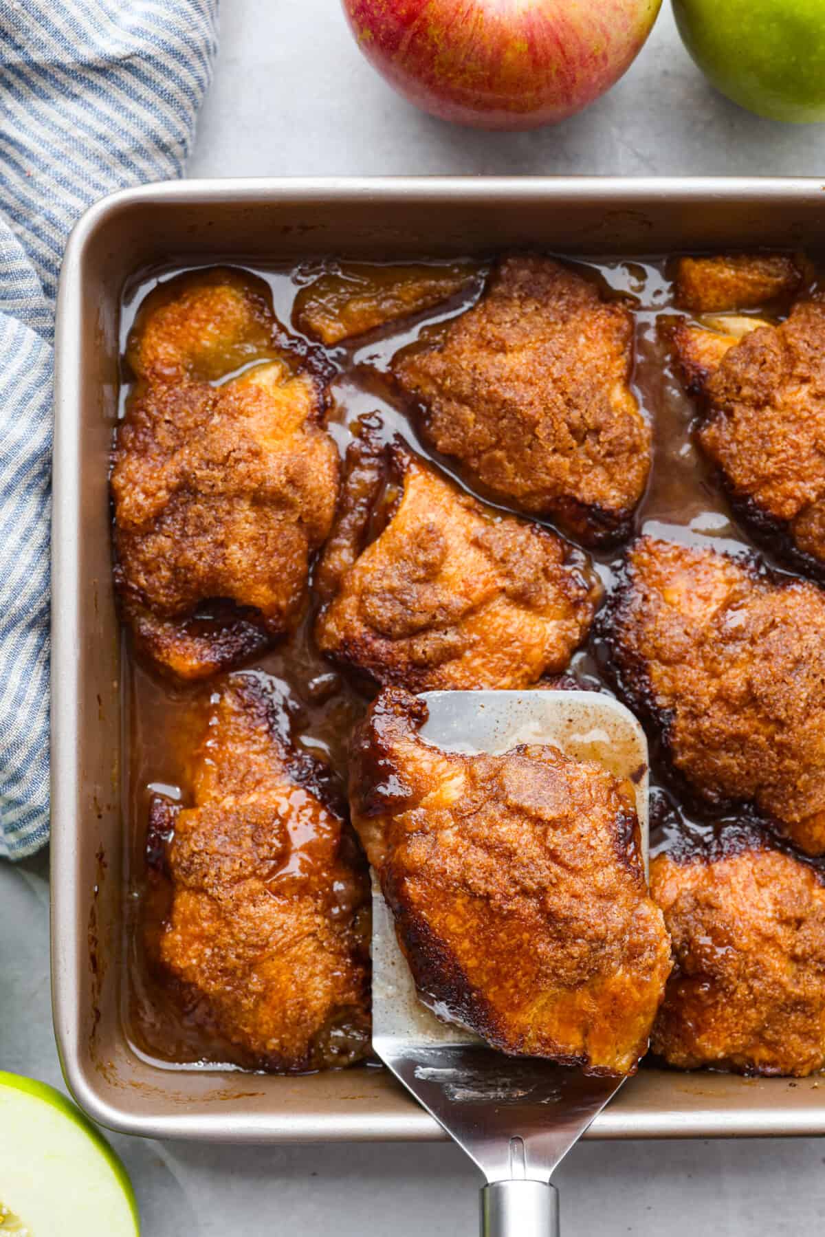 Top view of baked apple dumplings in a pan with a spatula. Apples are next to the pan with a striped kitchen towel.