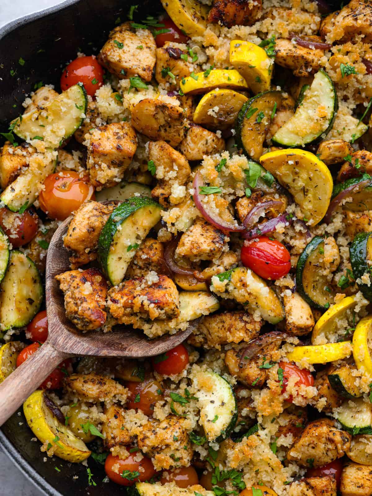 Close up view of a wooden spoon lifting up the chicken and garden vegetables from the skillet.