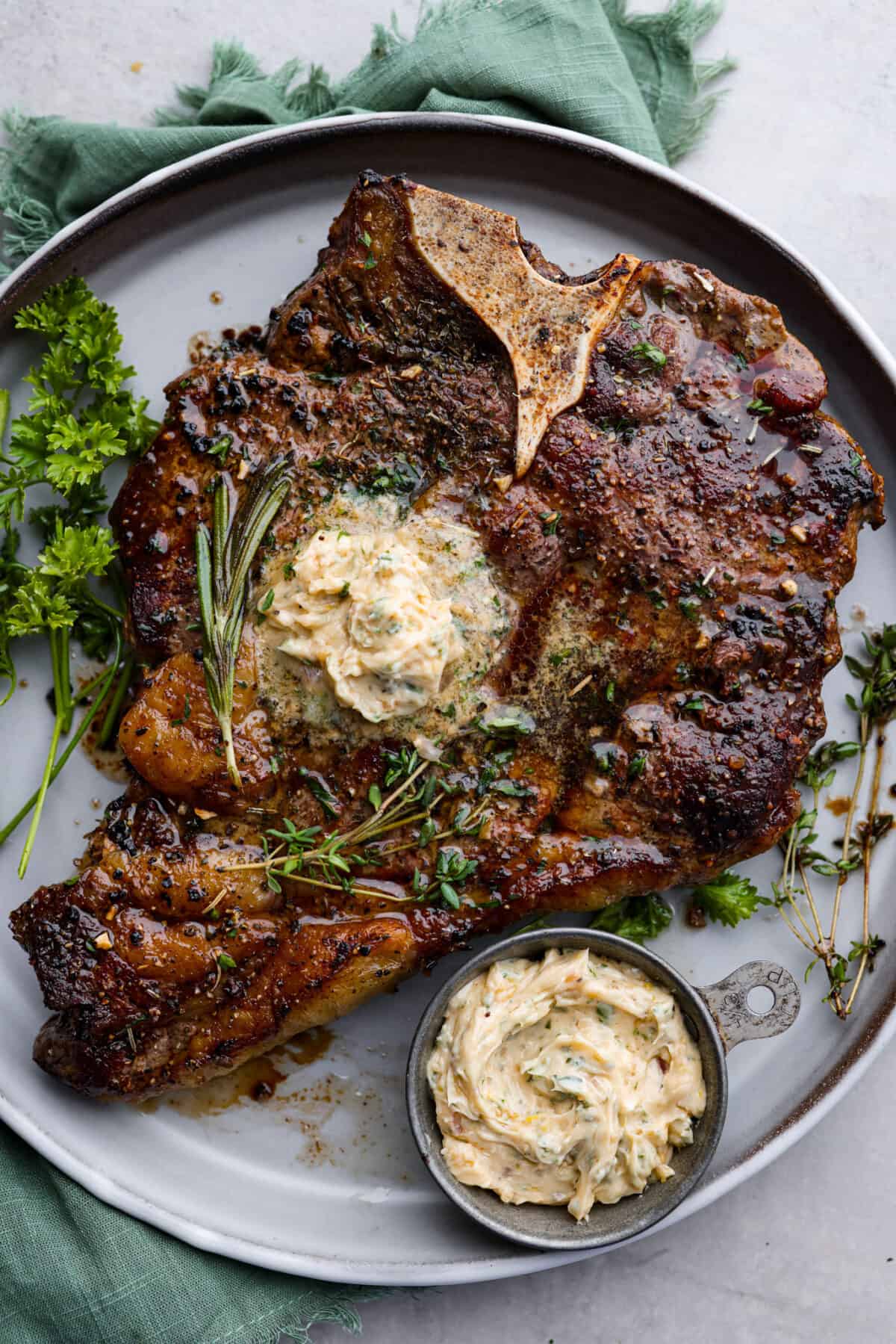 Top view of cowboy butter melted on top of a steak. A bowl of cowboy butter is next to the steak on a gray plate.